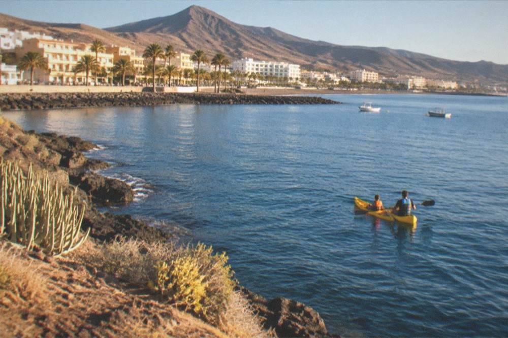 A child in a life jacket sitting in a kayak beside an adult on calm water off Los Cristianos off Los Cristianos