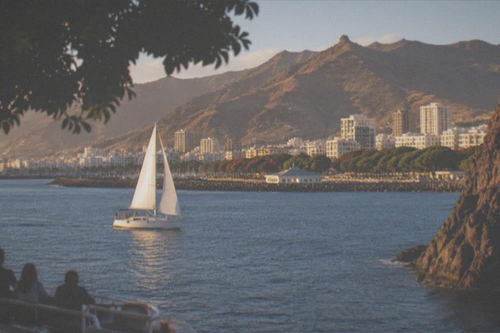 A sailboat on calm blue water near tall dark cliffs with people watching from the deck