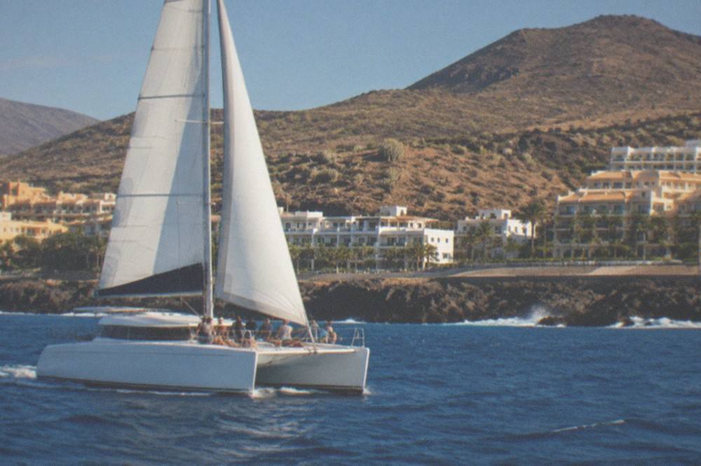 A catamaran under sail seen from the water with passengers on the foredeck
