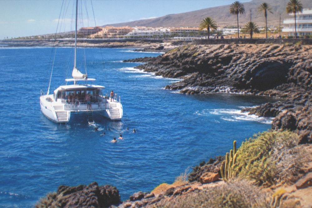 A catamaran at anchor in a rocky cove with passengers swimming near the stern