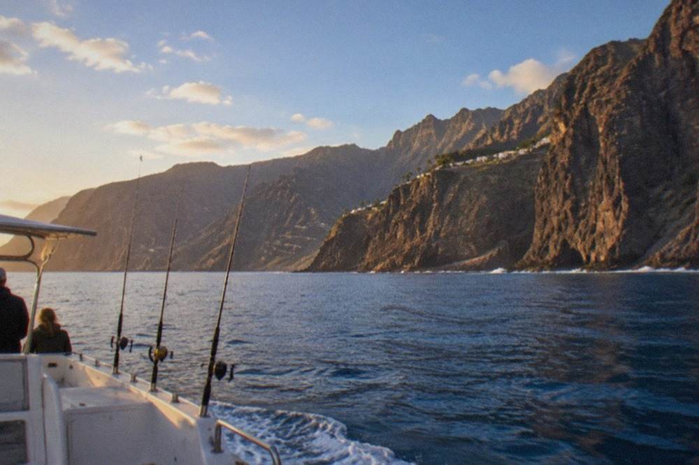 A small charter boat riding on calm blue water near the Tenerife coast with rod holders along the stern