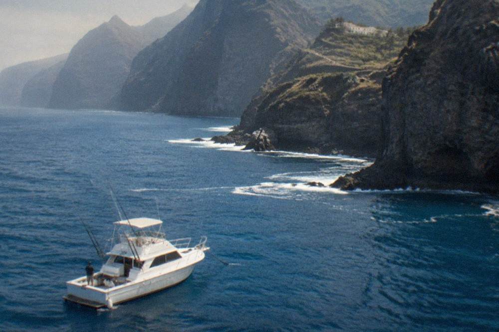 A charter boat anchored near tall dark cliffs with fishing rods resting in holders