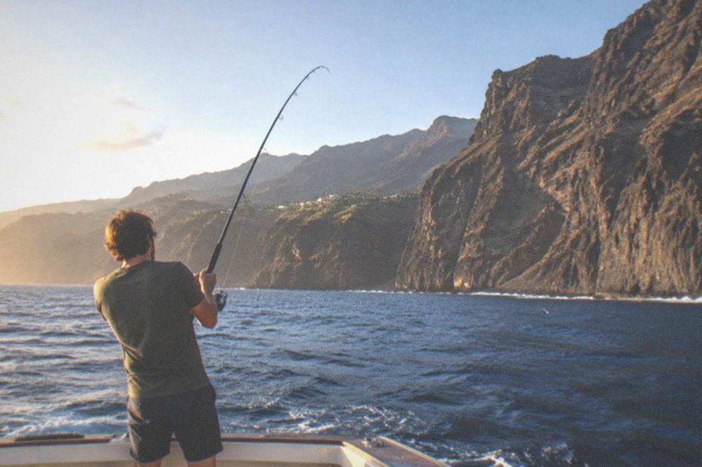 A person in shorts reeling in a fishing line from the stern of a boat on a sunny day