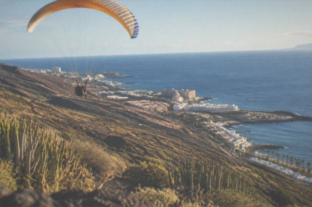 A tandem paraglider soaring above green hills near the coast