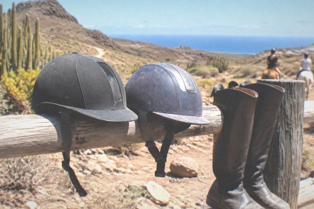 A pair of riding helmets and boots resting on a fence rail under bright sunshine