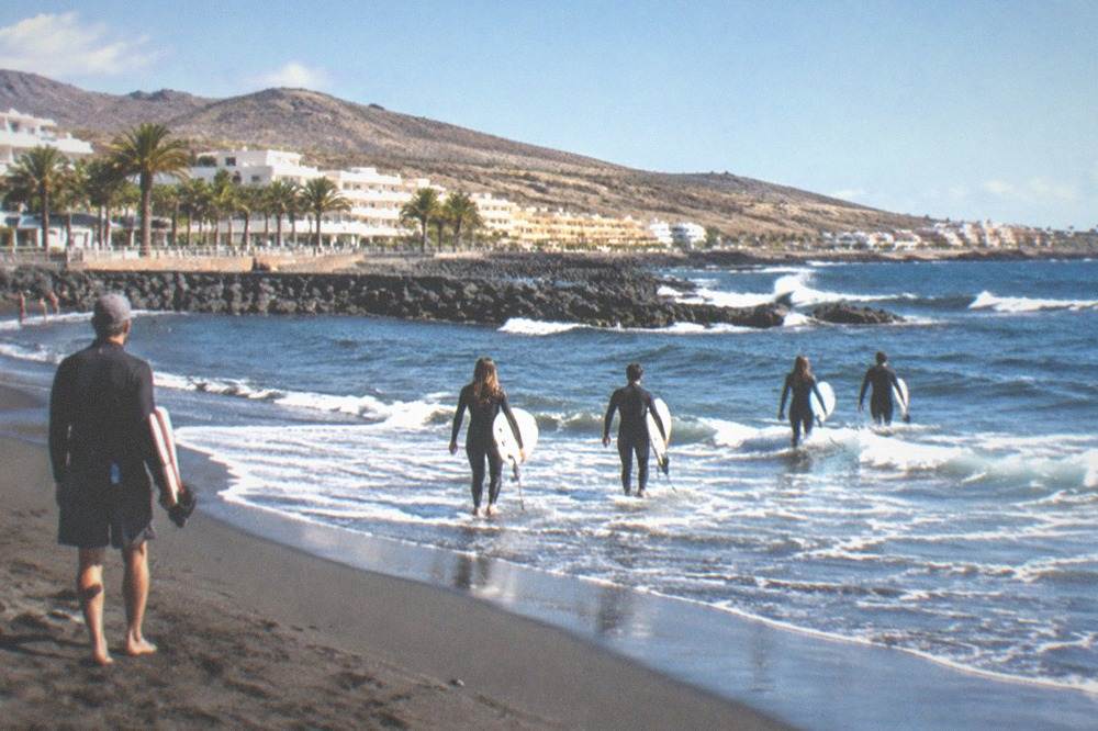 A group of surfers walking into small waves at a dark sand beach with a coach nearby