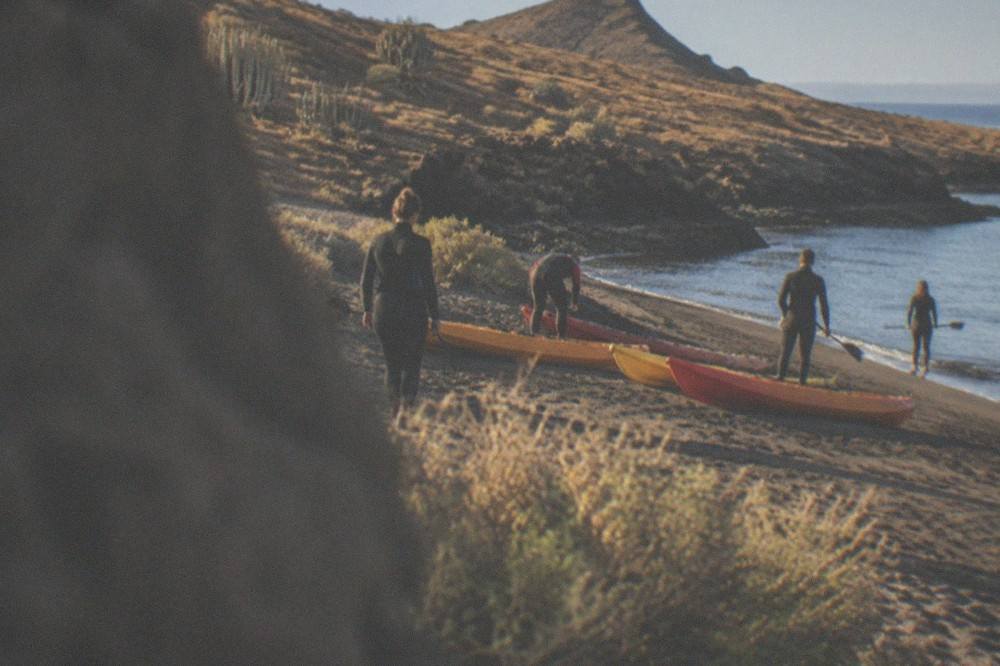A group of people in wetsuits preparing kayaks on a sandy beach beside calm water