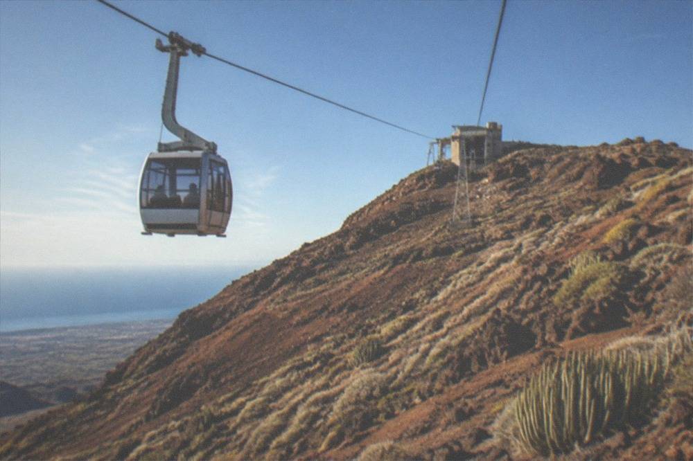 A cable car cabin ascending a barren red volcanic slope towards a station on a mountainside