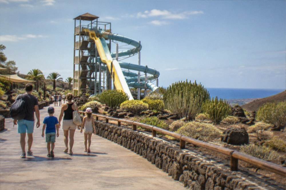 A family walking along a wide promenade towards a tall water slide tower in sunshine