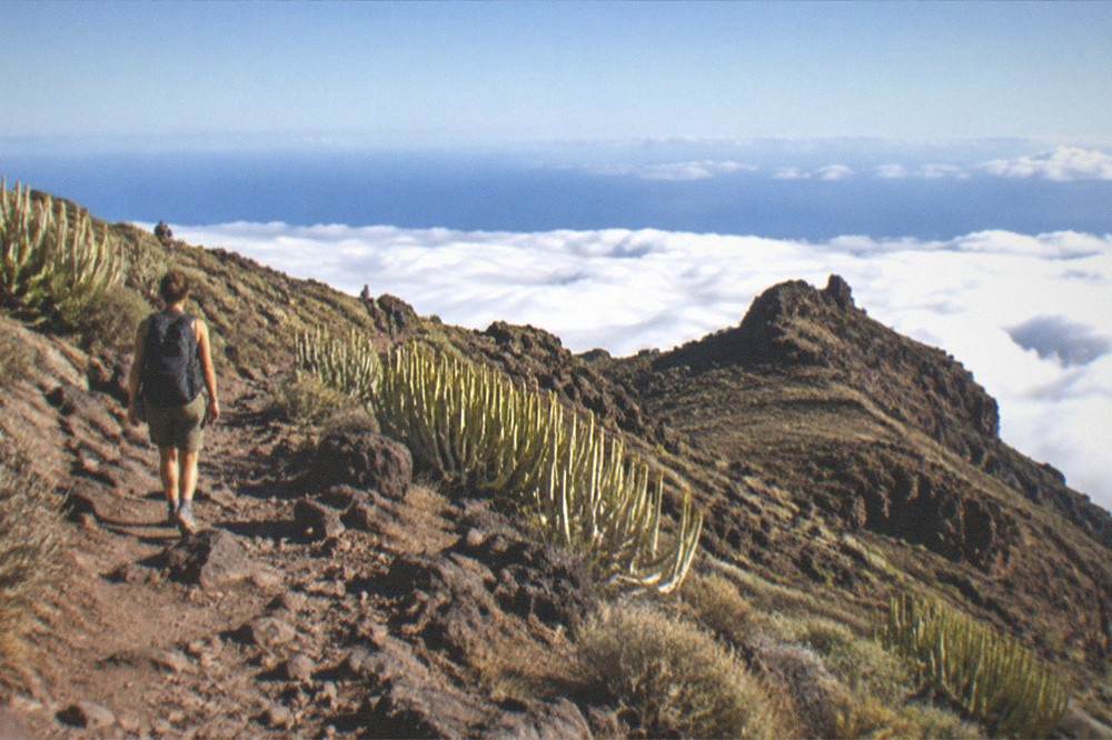 A hiker with a backpack walking a rocky path above a sea of clouds on a volcanic ridge