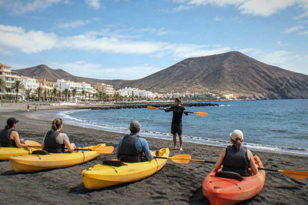 A guide demonstrating paddle technique to a small group seated in kayaks on a beach