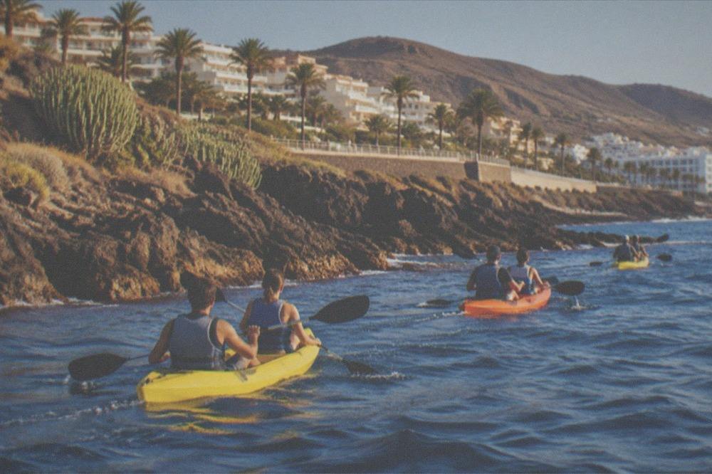 A group of double kayaks paddling in a line along a rocky coastline in morning light