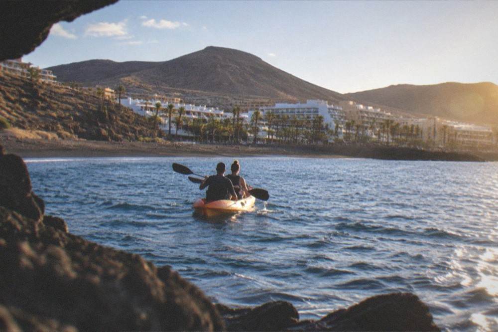 Two people in a double kayak seen from behind with a palm-lined beach in the distance