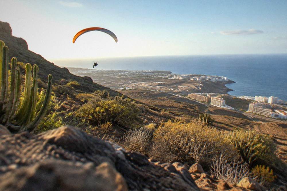 A paraglider flying along a rocky coastline with hotels visible on the shore below