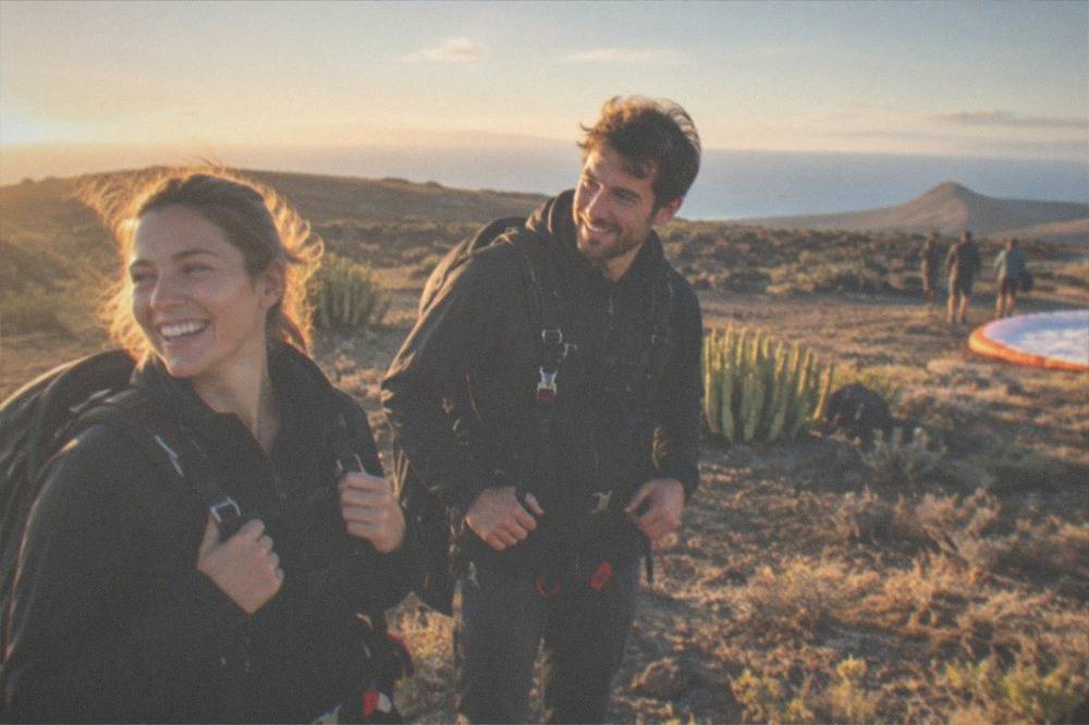 A passenger and pilot in matching harnesses smiling after landing on a coastal field