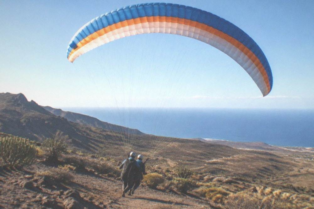 A paraglider wing fully inflated overhead with the passenger and pilot running down a launch slope