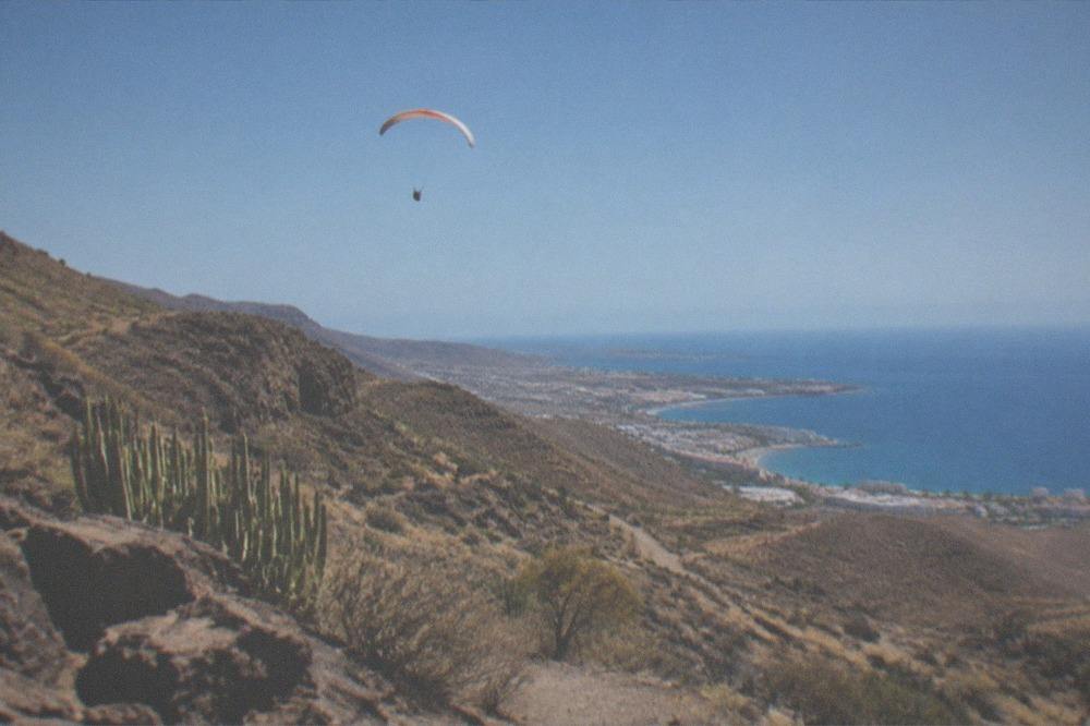A paraglider high above turquoise water with the Tenerife coastline curving into the distance