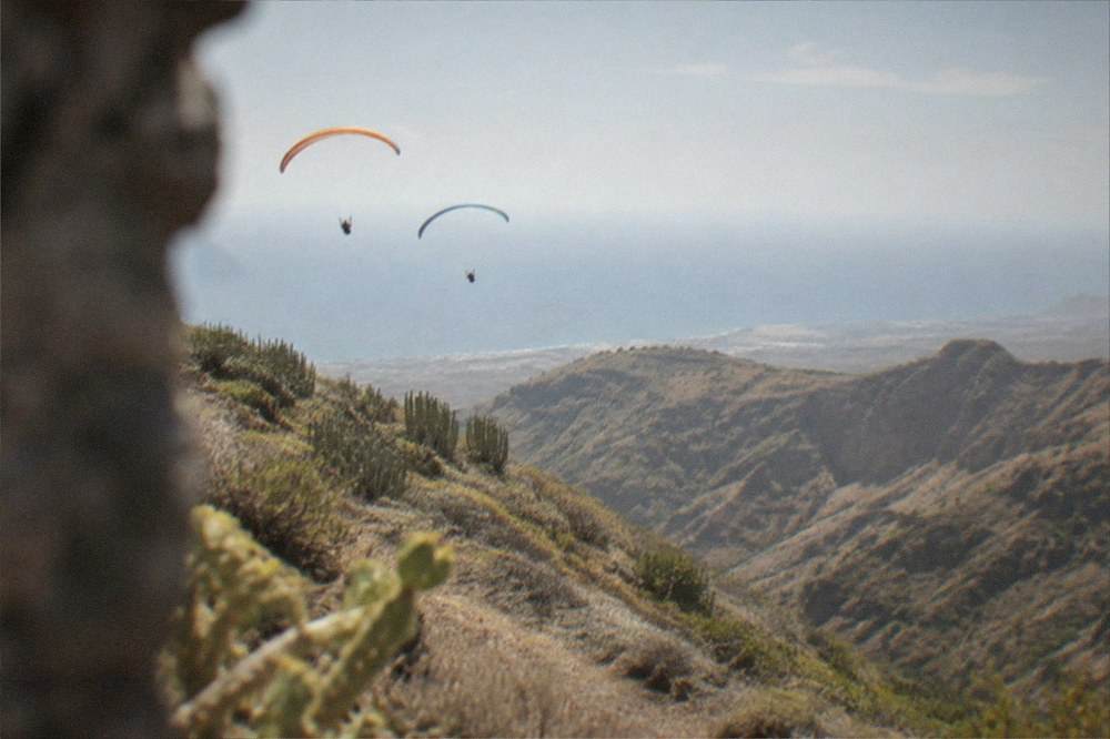 Two paraglider canopies flying in formation above green ravines near the coast