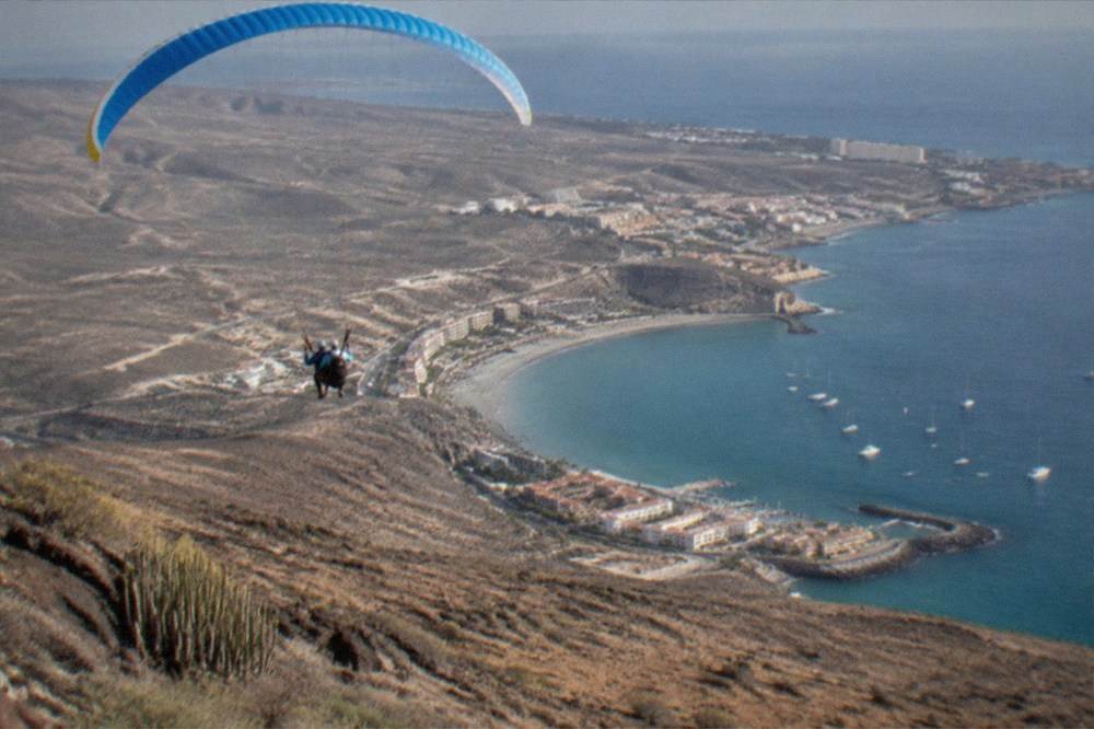 A tandem paraglider seen from above, flying over a bay with small boats anchored offshore