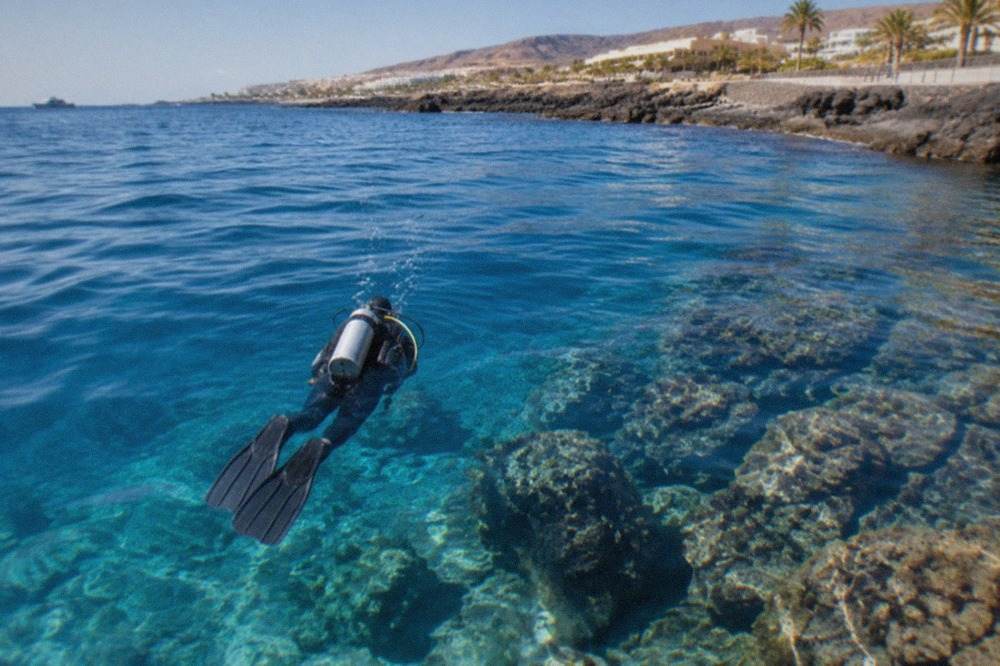A diver in a wetsuit swimming over a rocky volcanic seabed in clear blue water