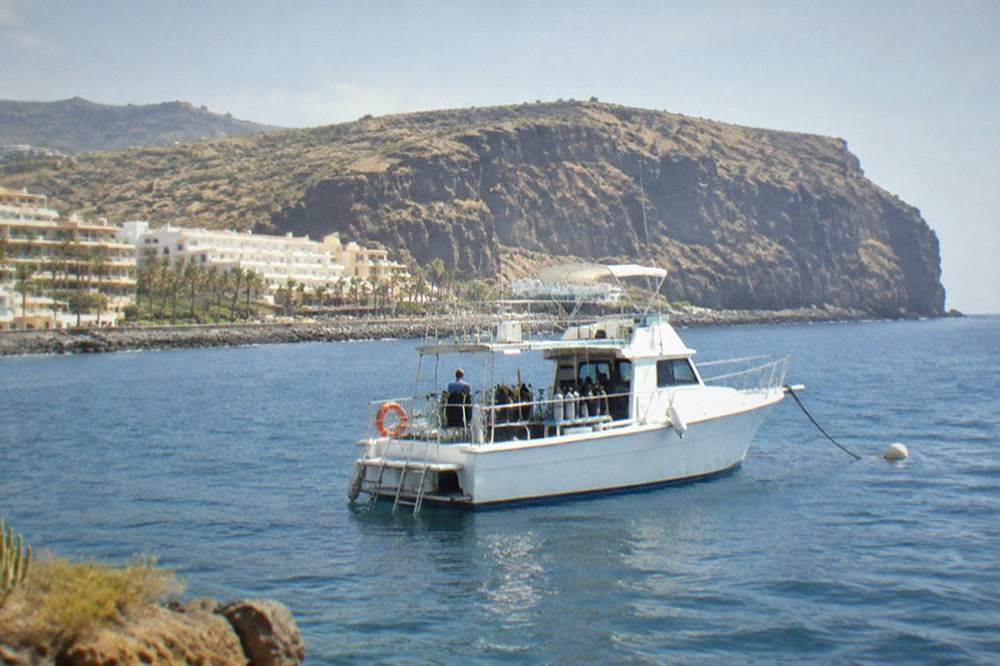 A dive boat anchored in a calm blue bay with a volcanic cliff behind it