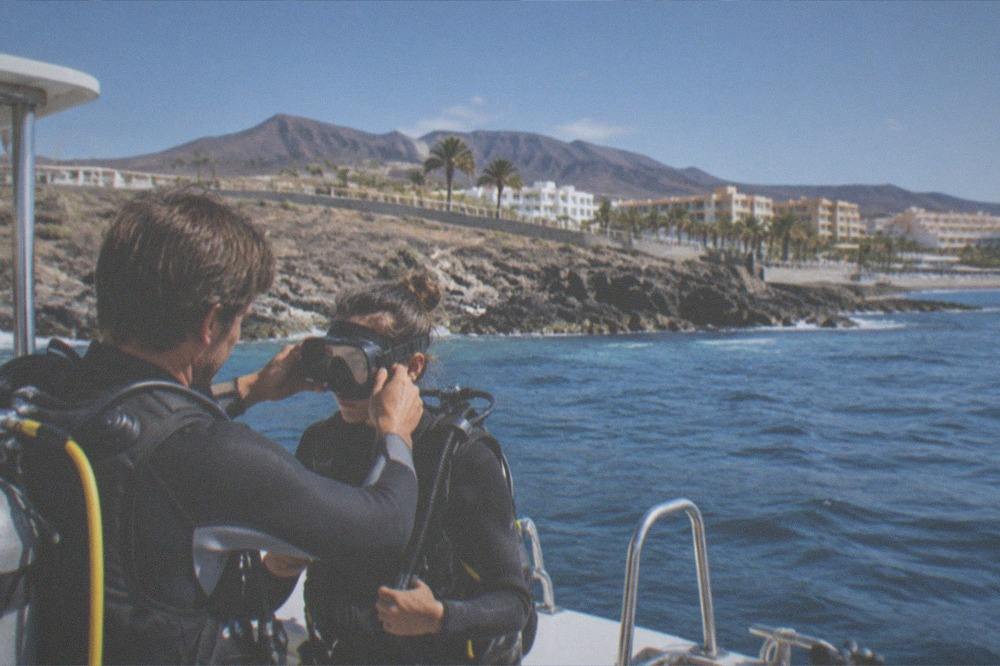 An instructor helping a beginner diver adjust a scuba mask beside a dive boat