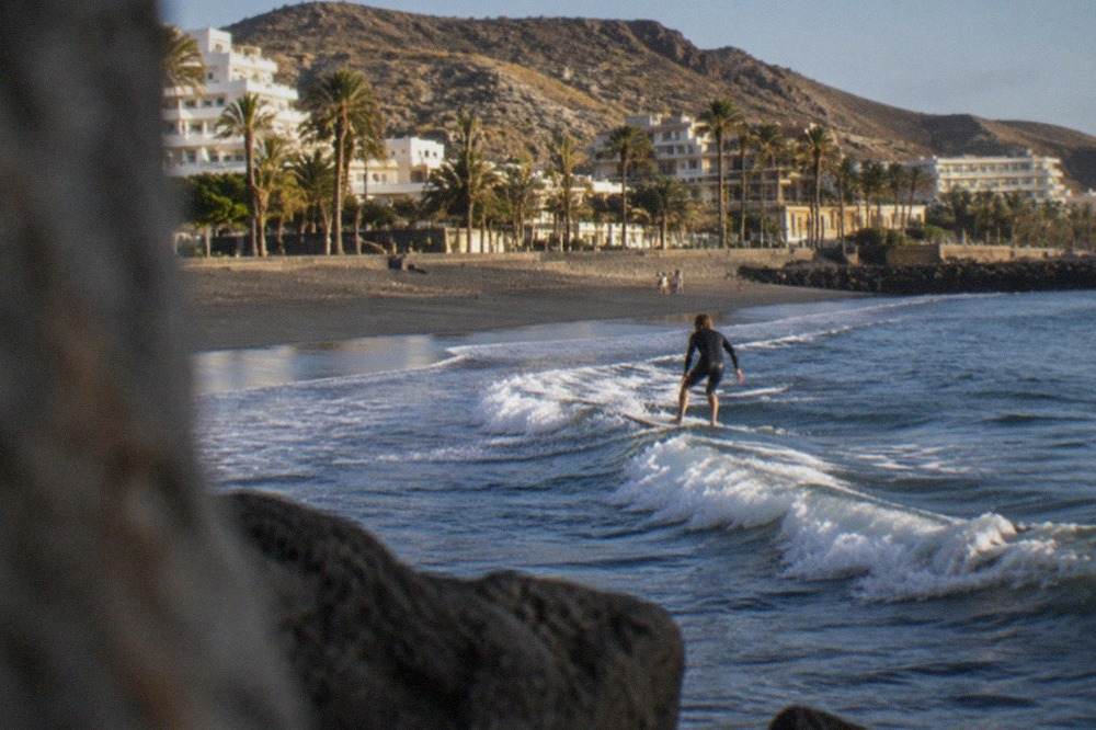 Surfer riding a small wave on a sandy beach with palm trees and hotel buildings behind