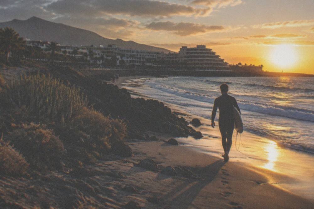 Sun setting over the Atlantic behind a silhouetted surfer walking with a board under one arm
