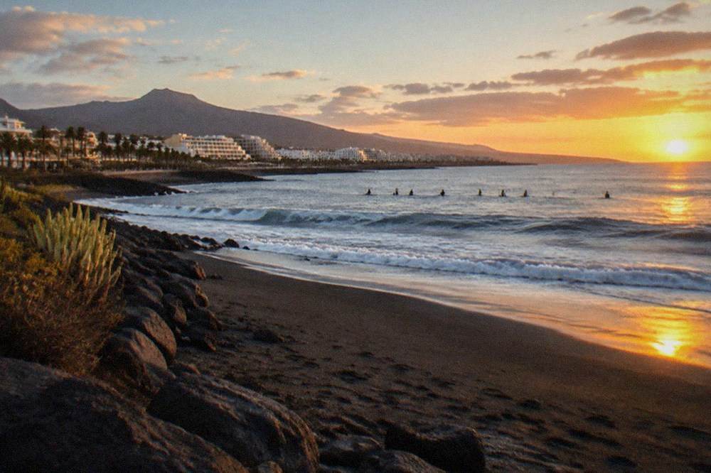Line of surfers sitting on boards beyond the break at sunrise on a volcanic sand beach