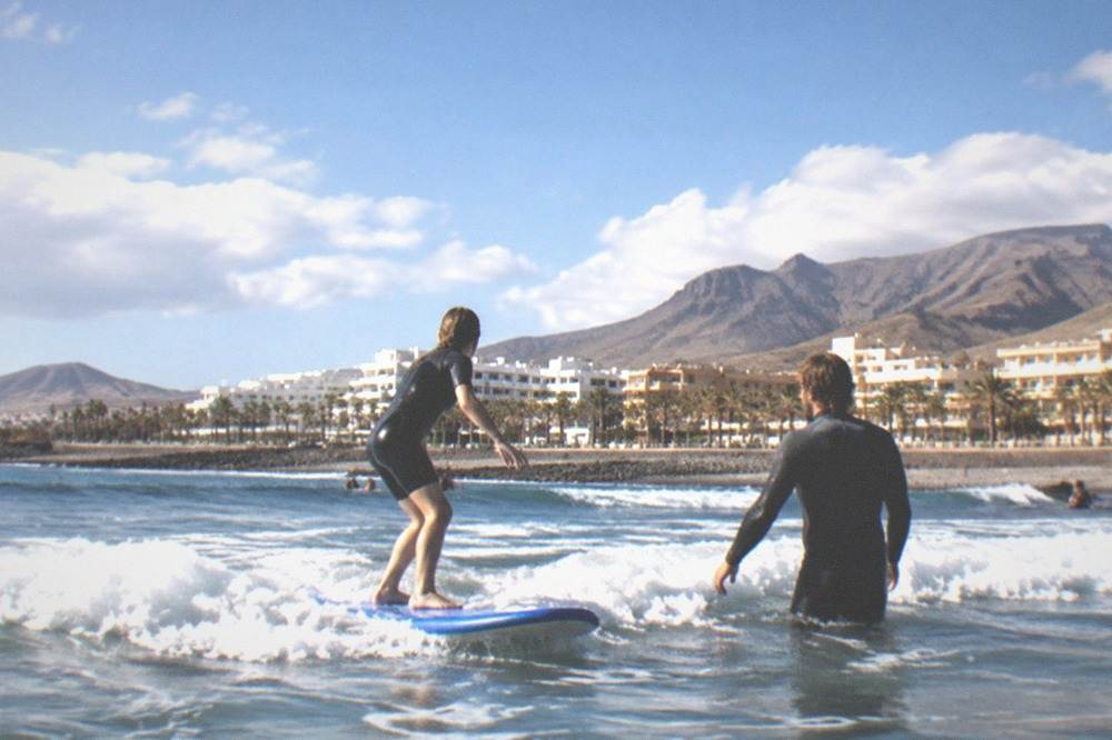 Learner surfer standing on a board in shallow whitewater with an instructor nearby