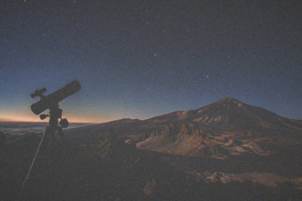 A telescope silhouetted against a starry night sky above dark volcanic ground