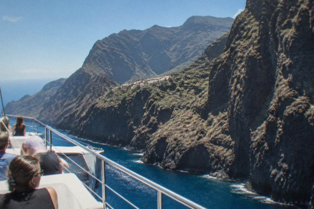 A catamaran with passengers sailing along the coastline past tall dark cliffs