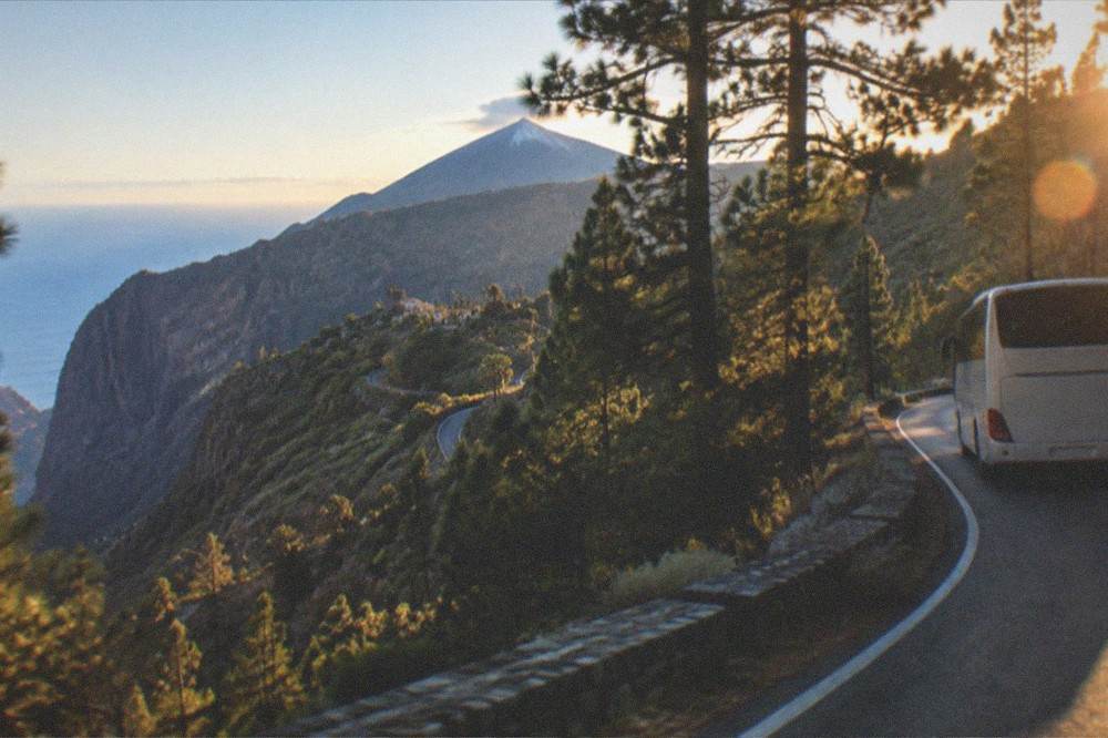 A tour coach driving a winding mountain road through pine forest with a volcano in the distance