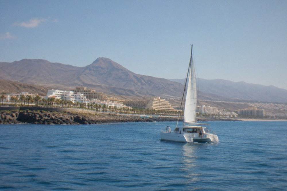 A small catamaran cruising on calm blue water off the Tenerife coast with hills in the distance