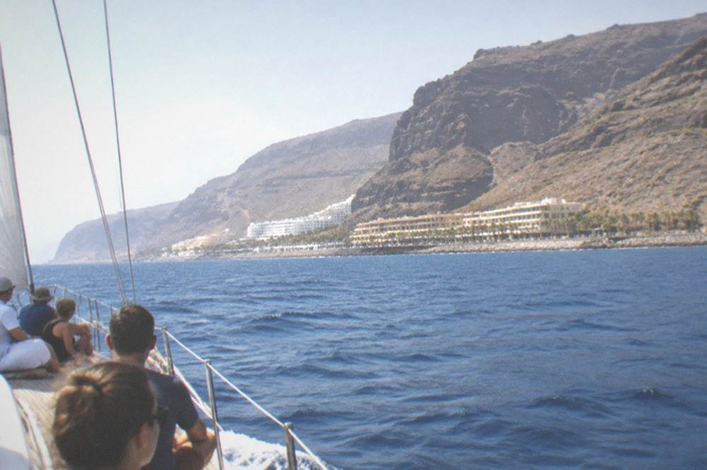 A sailboat under way near tall cliffs with passengers seated along the deck